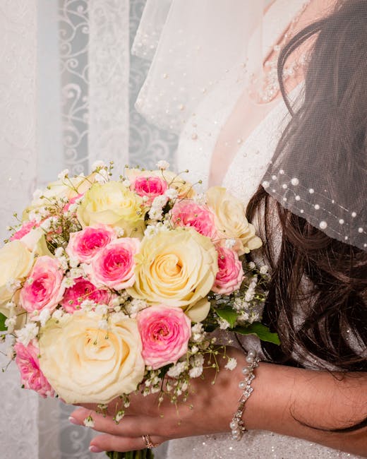 A close-up of a bride holding a bouquet of fresh flowers, featuring large cream-colored roses and smaller pink spray roses, accented with delicate white baby's breath. The bouquet is wrapped in white 