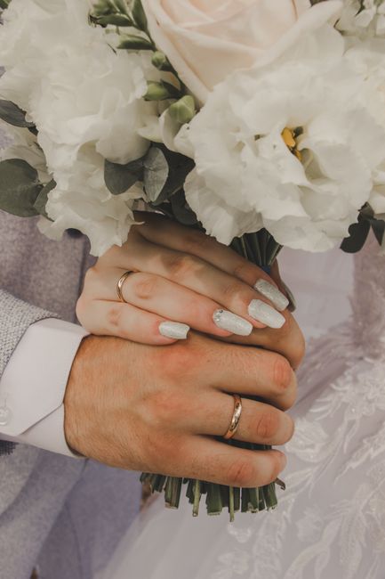 Close-up of a newlywed couple holding a bouquet of fresh white lisianthus, roses, and eucalyptus with their hands intertwined. The bride's nails are painted with a shimmering silver polish, and she we
