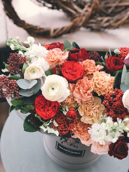 A vibrant floral arrangement featuring a variety of fresh flowers including red roses, white ranunculus, peach carnations, and dark red berries, all artistically arranged in a white cylindrical box. T