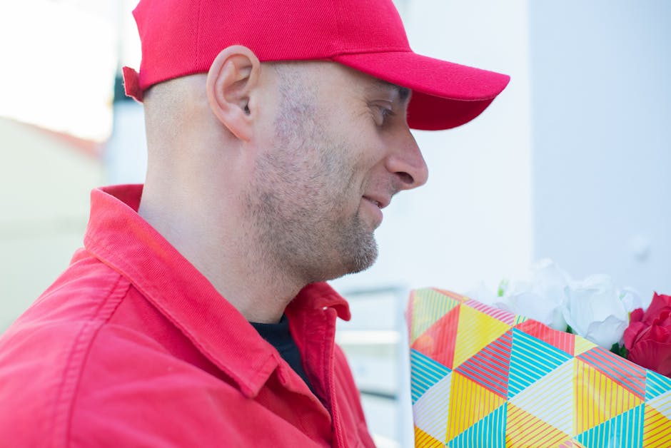 A man wearing a red cap and matching red jacket is holding a colorful floral gift wrapped in geometric patterned paper with vibrant shades of yellow, red, blue, and white. The bouquet contains white a