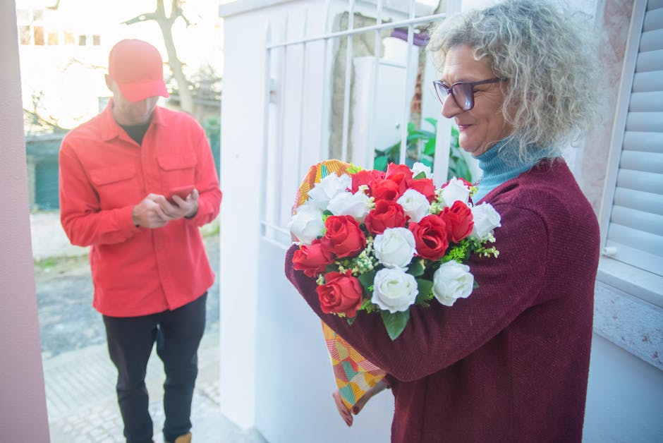 A woman with curly gray hair, wearing glasses, a maroon sweater, and a blue turtleneck, stands at a doorway holding a large, freshly arranged floral bouquet featuring red and white roses with green fo